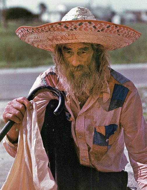 older weathered man in a large sombrero hat holding a cane. Lawnmower near by.